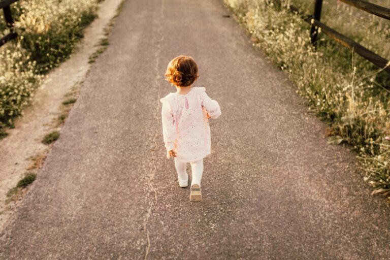 A young child in a pink dress walks alone on a rural path during sunset, surrounded by nature.
