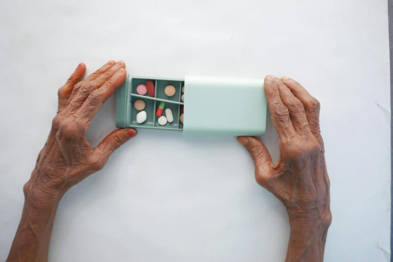 Elderly person sorting pills into a turquoise pillbox on a white surface.