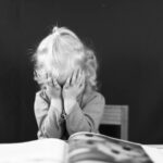 Black and white photo of a young child covering their face with hands, sitting in front of a book.