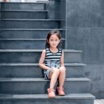 A young girl in a striped dress sits on a sleek grey staircase, looking into the camera.