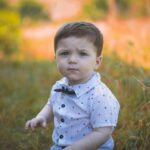 Adorable baby boy sitting outdoors in a sunny park, surrounded by vibrant grass and flowers.