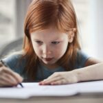 Focused young girl with red hair writing on paper at a desk indoors.