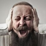 Portrait of a man with facial hair, covering his ears and screaming, expressing stress or frustration.