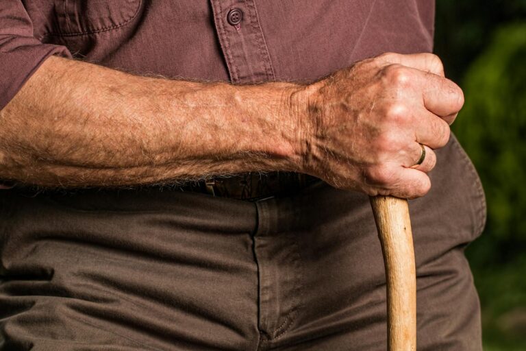 Close-up of a senior adult's hand gripping a wooden walking cane outdoors, symbolizing support and aging.