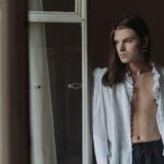 An elegant androgynous model poses by an indoor window, exuding subtle sophistication.