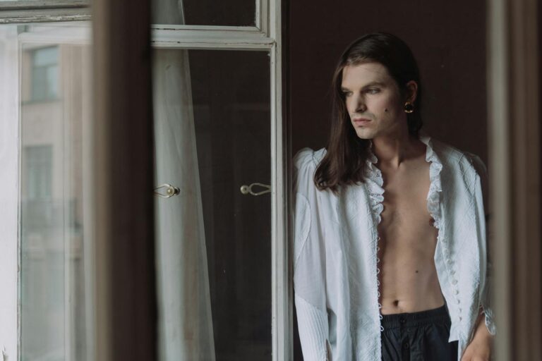 An elegant androgynous model poses by an indoor window, exuding subtle sophistication.