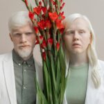 Charismatic couple in trendy outfits standing with bunch of red flowers with lush leaves and looking at camera while standing against beige background