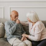 An elderly woman comforts a man coughing on a couch, showcasing care and affection.