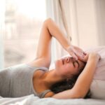 Woman in gray tank top resting on bed with natural light from window, looking relaxed and comfortable.