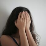 An adult woman with long brunette hair covers her face with her hands against a white wall, creating a minimalist portrait.
