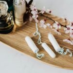 Flat lay of feminine hygiene products, perfumes, and cherry blossoms on a wooden tray.