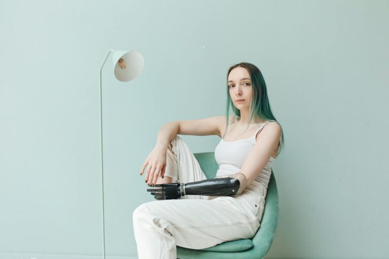 Portrait of a woman with prosthetic arm seated in a stylish interior, wearing a white tank top.