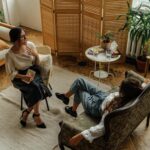 Two women engaged in a psychotherapy session in a warm, inviting interior with plants and natural lighting.