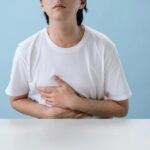 An adult man in a white shirt holds his stomach, indicating pain and discomfort, with a light blue background.