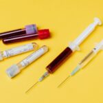 From above of medical syringe with medication near injector with blood sample arranged with filled clinical test tubes placed on yellow background