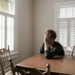 A man sits alone at a table in a bright room, displaying deep contemplation.